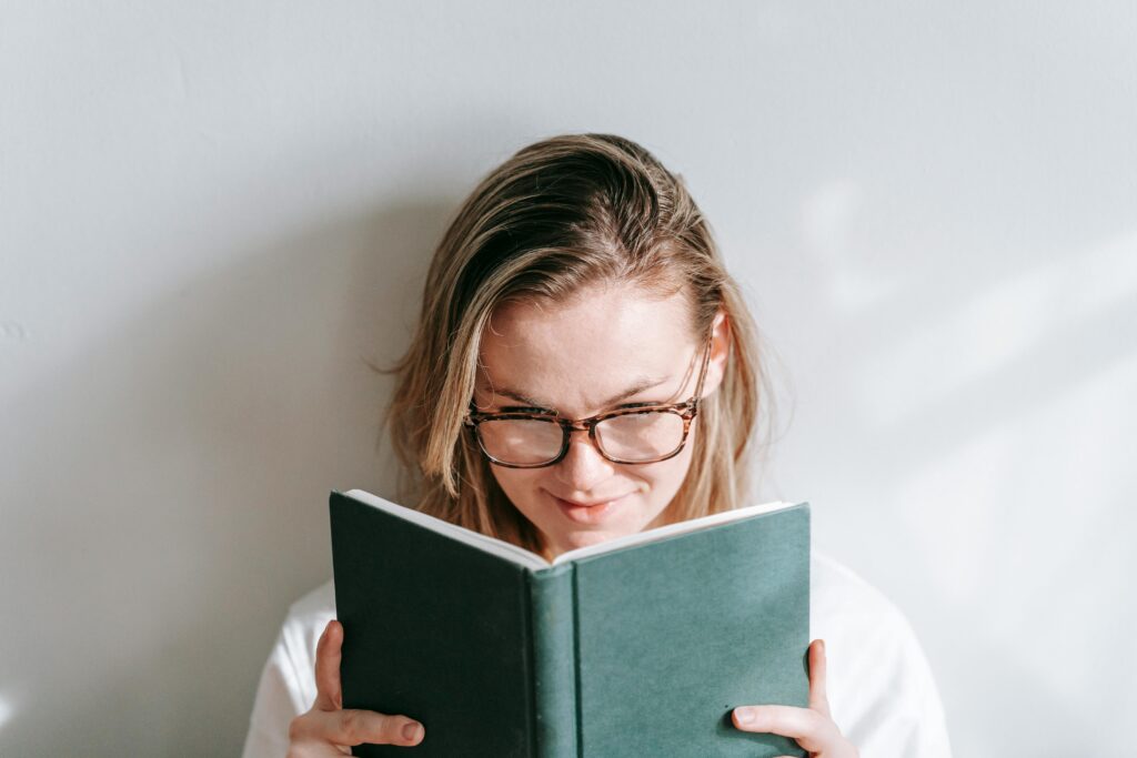 Interested young woman in glasses reading textbook while studying in sunshine on light background